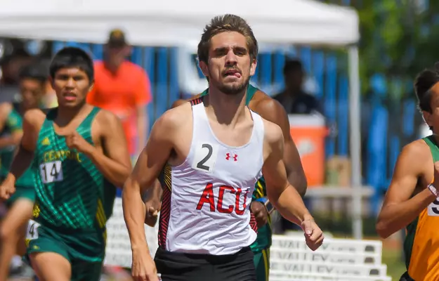Male sprinter in a white ACU singlet with number 2 leads a crowded outdoor track race, behind him are runners in green uniforms on a sunny day.