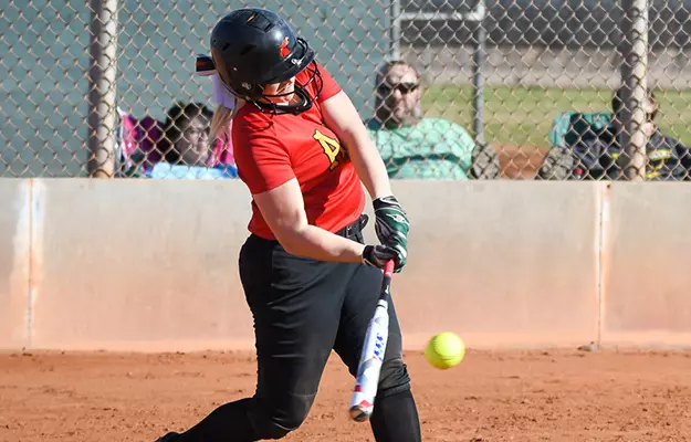 Female softball batter in a red jersey swings at a pitch on a dirt field, helmet on, spectators behind the fence.