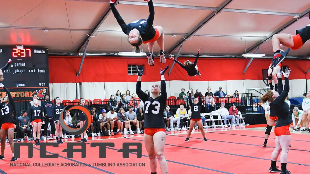 Cheerleaders perform a high basket toss in a red gym while spectators watch from the bleachers, with a large NCATA logo on the floor.