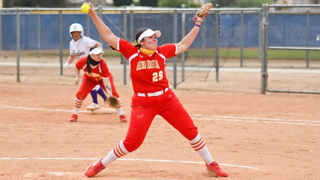 Arizona Christian softball pitcher in red, #29, mid-pitch on a dirt field with a yellow ball raised in her throwing hand