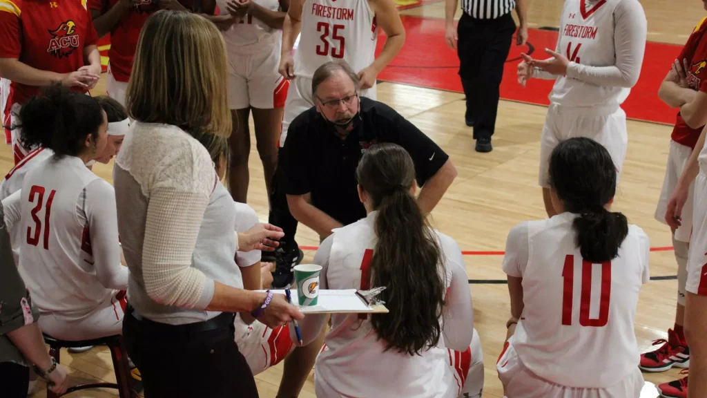 Basketball coach crouches and speaks to his white-jersey team during a huddle on the wooden gym floor, players gathered around a clipboard and notes.