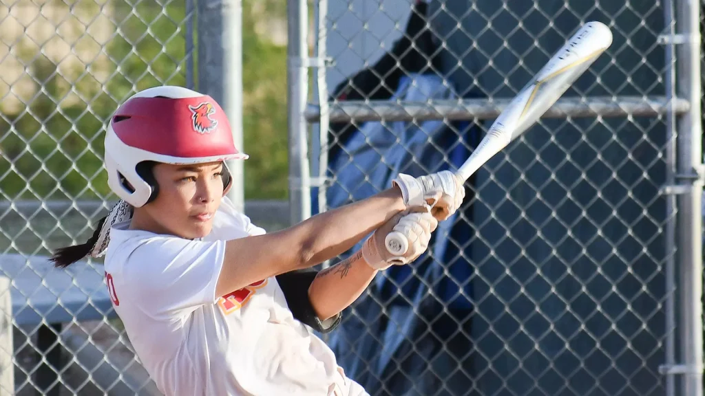Female softball player in a white uniform swings a bat, wearing a red helmet with a bird logo, near a chain-link fence.