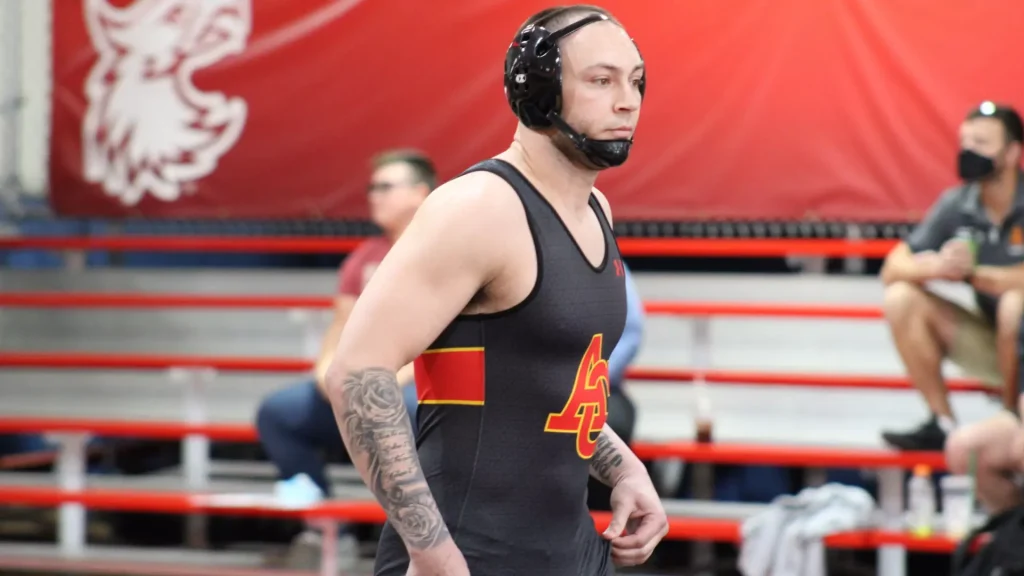 Wrestler in a black singlet with red and yellow design, wearing headgear, standing in a gym with red ropes in the background.