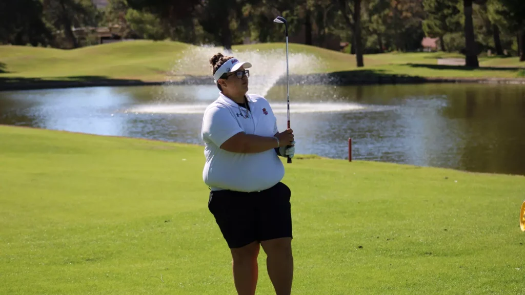Woman golfer in a white polo and visor stands on a green by a lake, holding a golf club ready to swing.