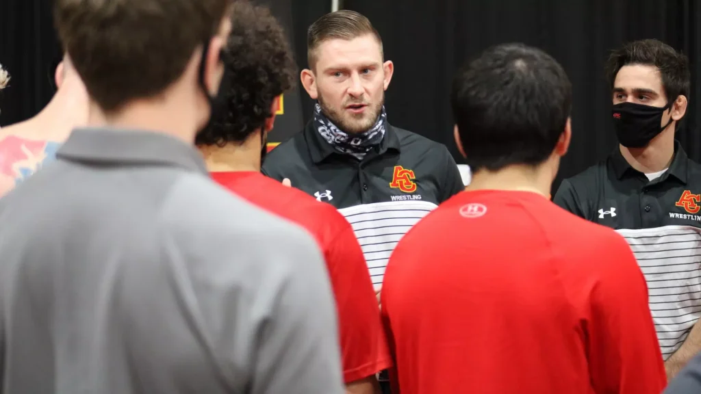 Coach wearing a black polo with a wrestling logo speaks to a circle of wrestlers in red jerseys, in a gym setting.