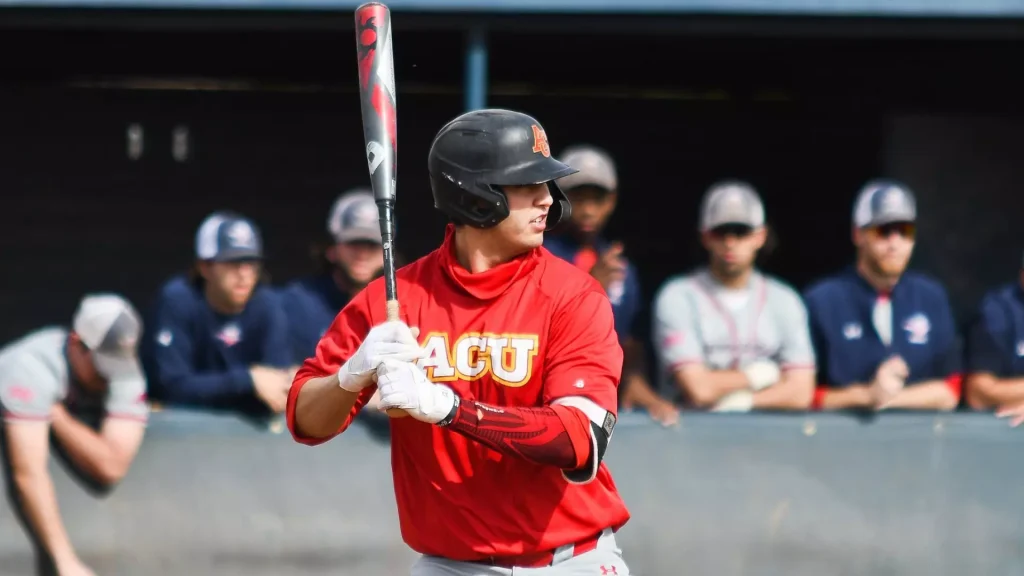 Baseball batter in a red uniform with helmet and white gloves, preparing to swing at the plate with teammates blurred in the background.