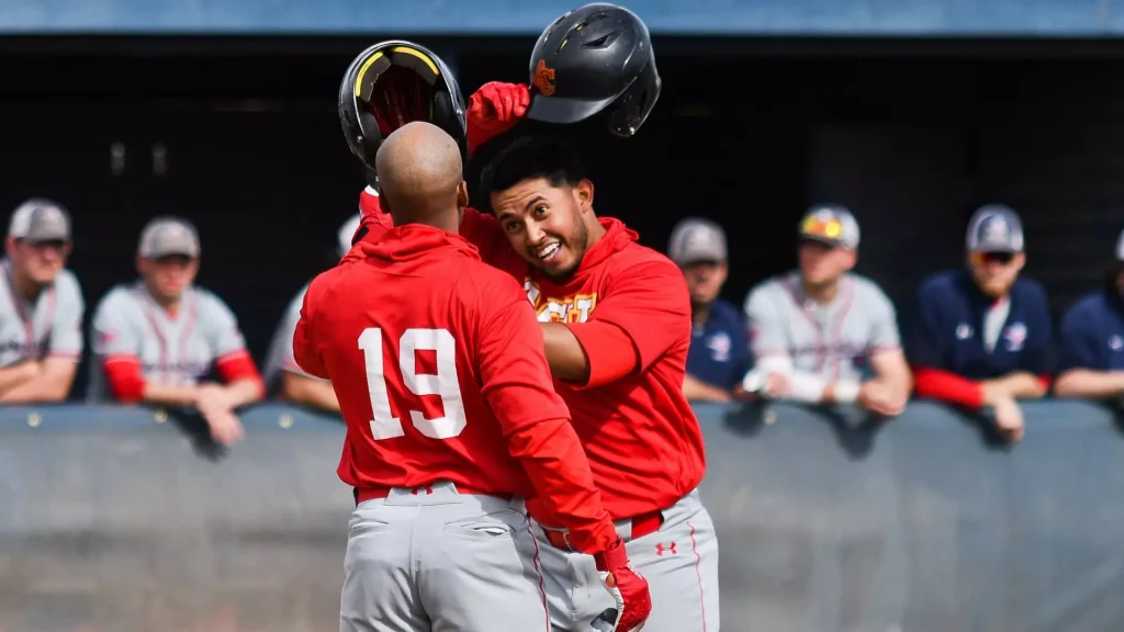 Two baseball players in red uniforms celebrate, lifting helmets, as teammates watch from the dugout in the background.