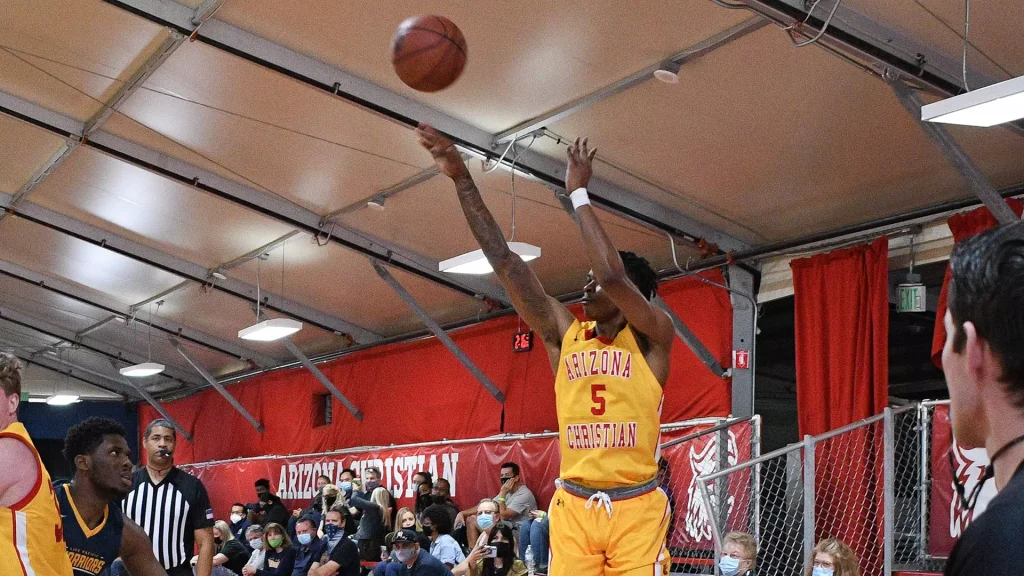 Basketball player in a yellow Arizona Christian jersey (No. 5) leaps to take a shot as a crowd watches indoors.