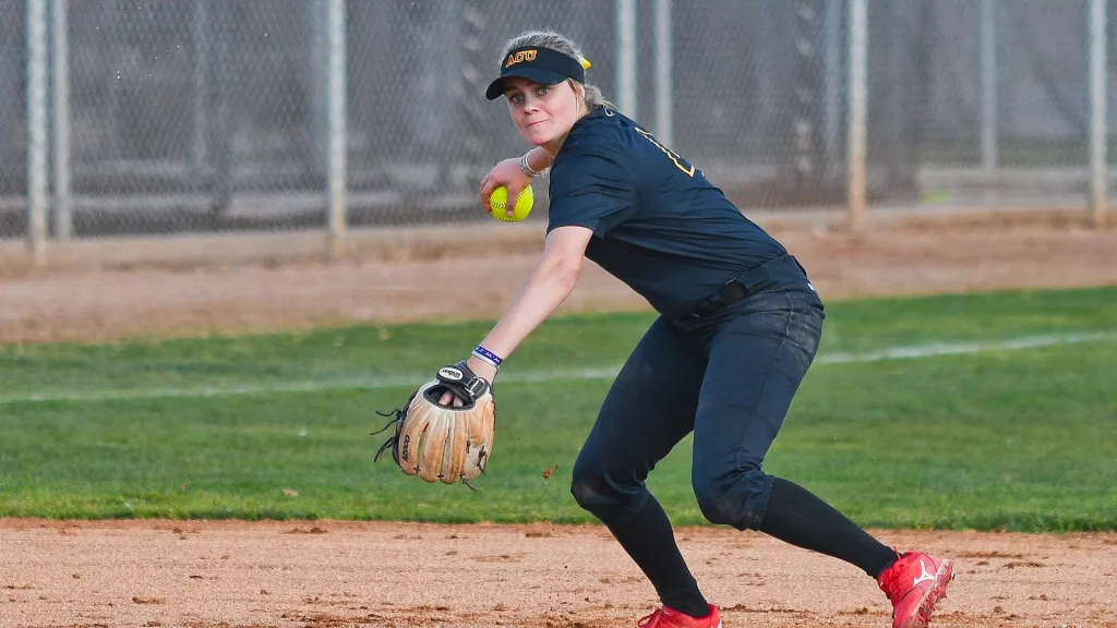 Female softball pitcher in a navy uniform mid-pitch on dirt infield, glove ready and ball in hand.