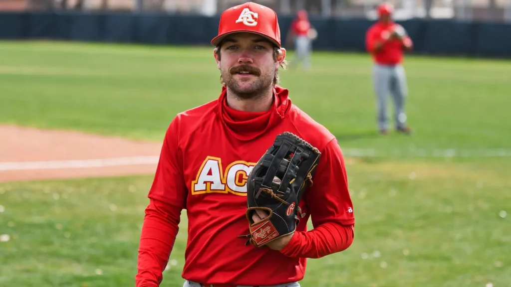 Baseball player in a red uniform and cap holding a glove on a grassy field with teammates blurred in the background.