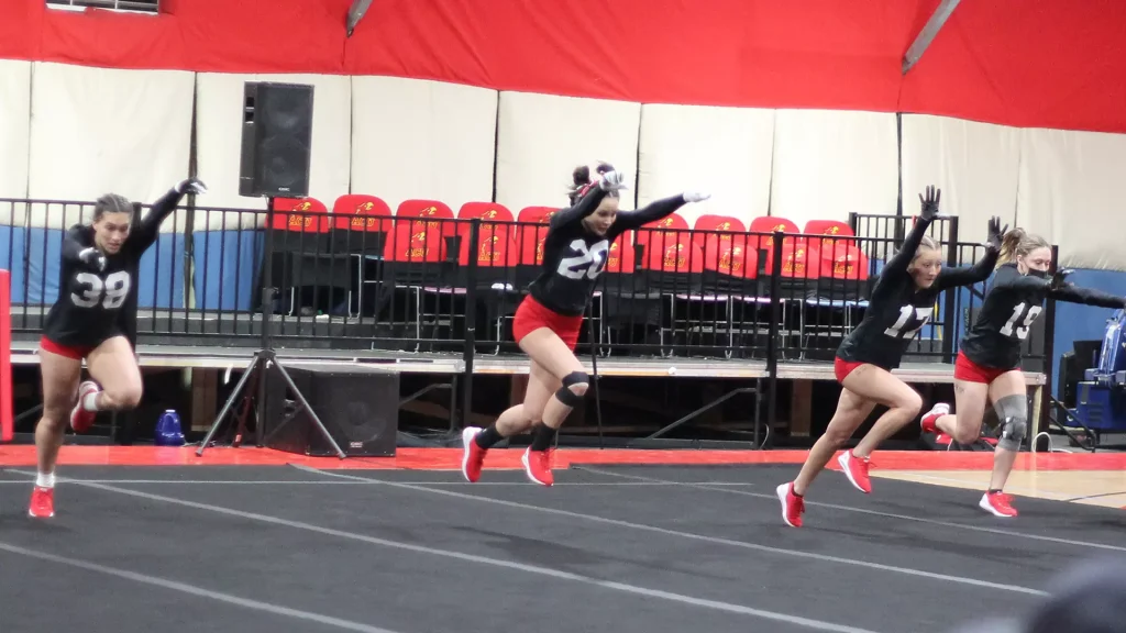 Four female sprinters in black jerseys and red shorts race on an indoor track, numbers 39, 20, 14, and 11 visible, with red arena seats in the background.