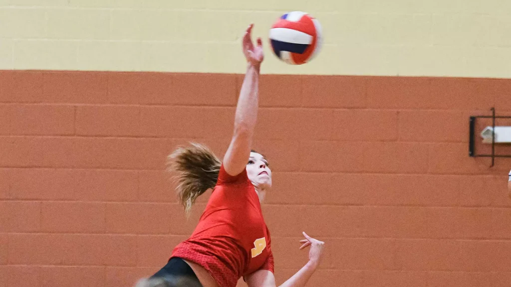 Female volleyball player in red jumps to spike a ball in an indoor gym, her arm extended toward a volleyball against an orange wall.