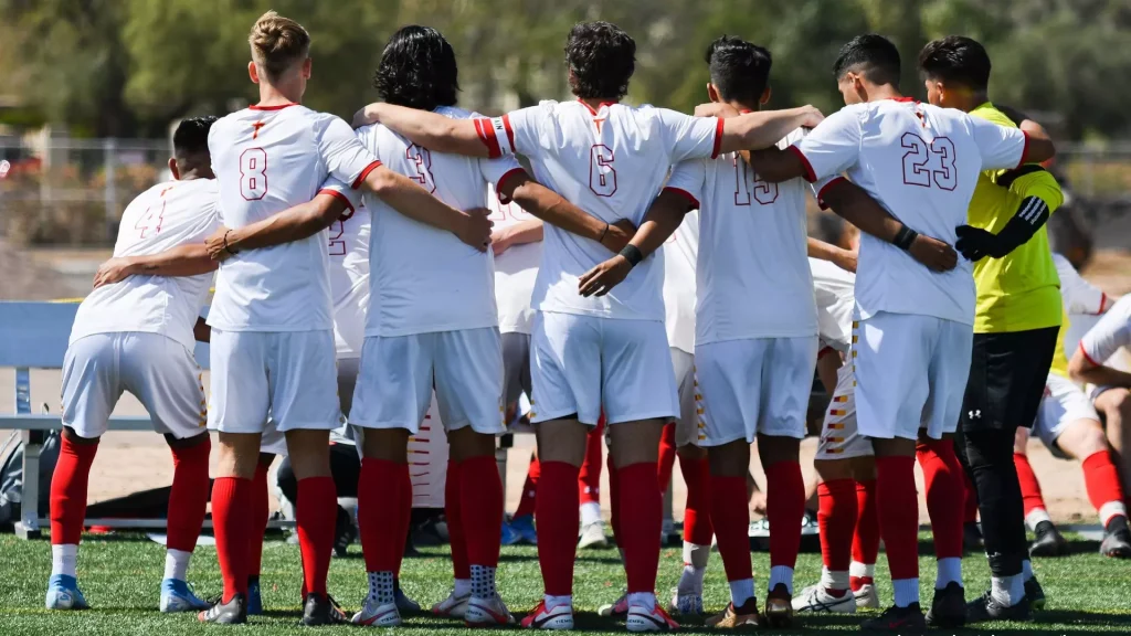 Soccer players in white uniforms with red trim stand in a line with arms around each other's shoulders on a green field, forming a huddle before the game.