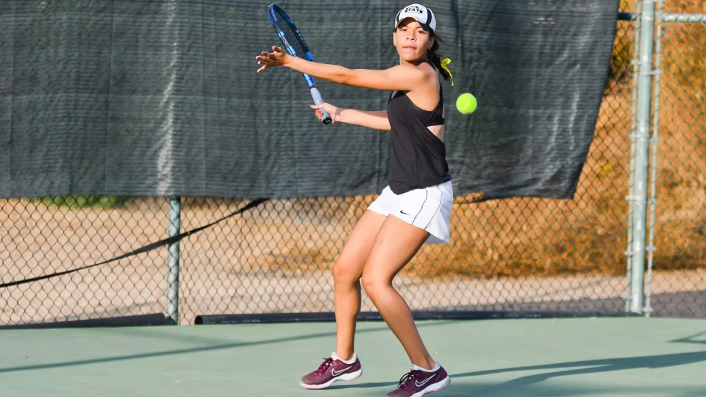 Female tennis player in a black tank top and white shorts taking a forehand on an outdoor court, wearing a cap and maroon sneakers