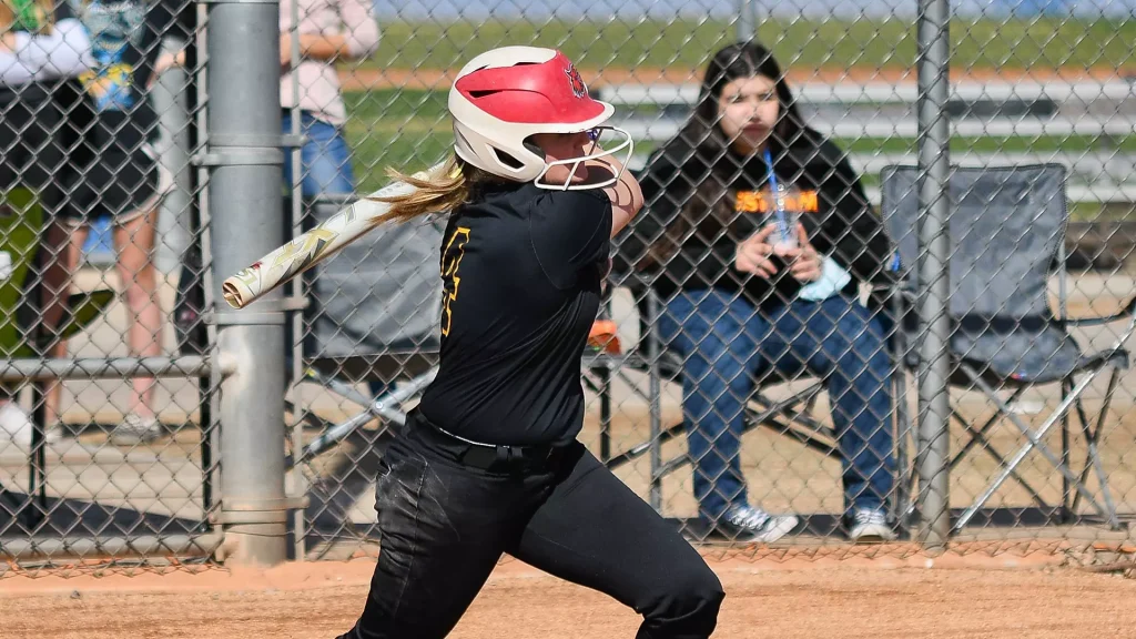 Female softball player in a black uniform swings a bat at the plate, wearing a red-and-white helmet behind a chain-link fence.
