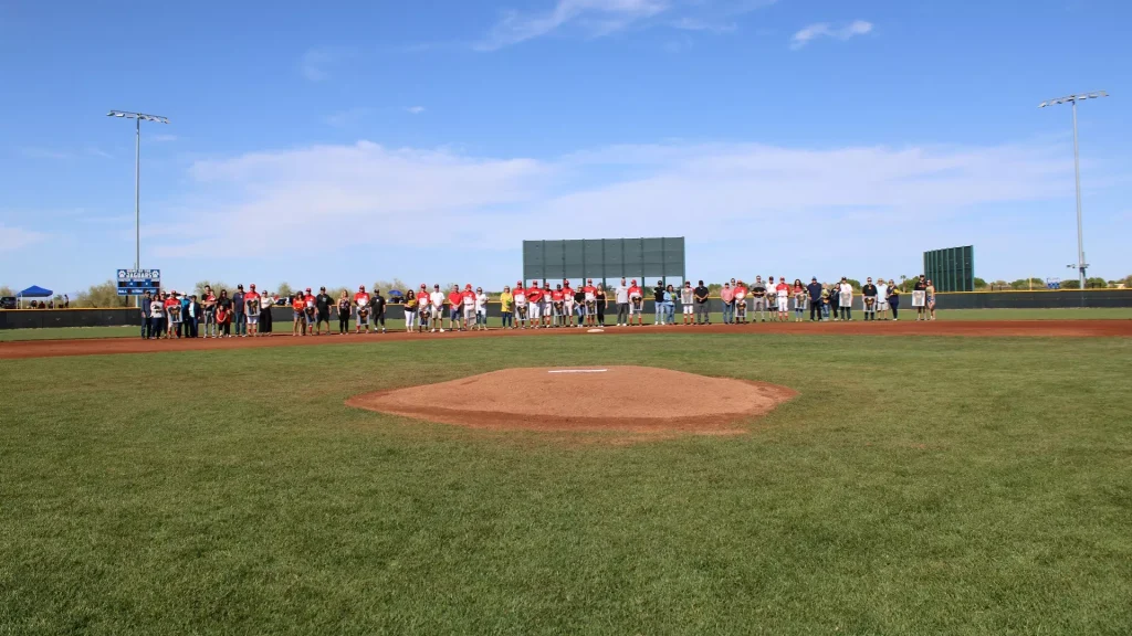 A large group of people standing along the baseline of a baseball field during a ceremony, with the pitcher's mound in the foreground and a clear blue sky overhead.