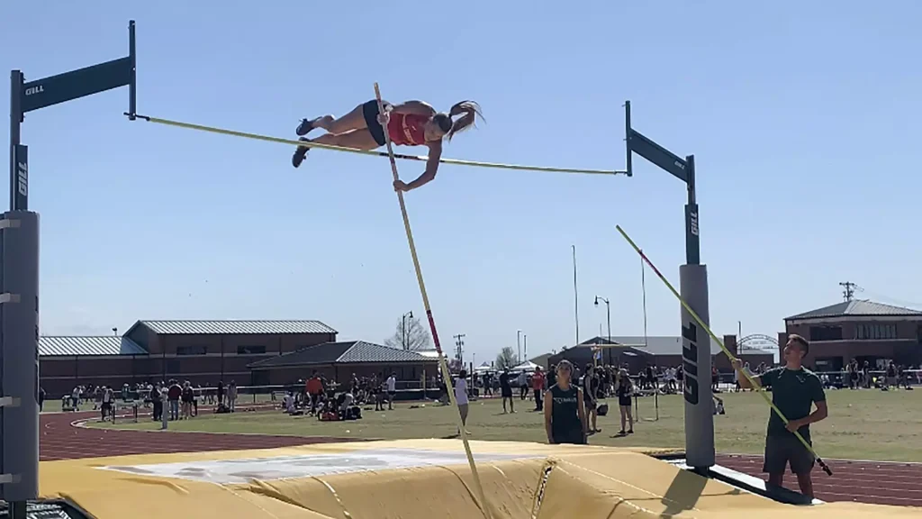 Female pole vaulter in a red top clears a horizontal bar outdoors during a track and field meet.