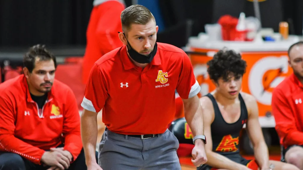 Male wrestler in a red polo with a team logo leans forward on the mat during a match, focused and tense, with teammates seated behind him.