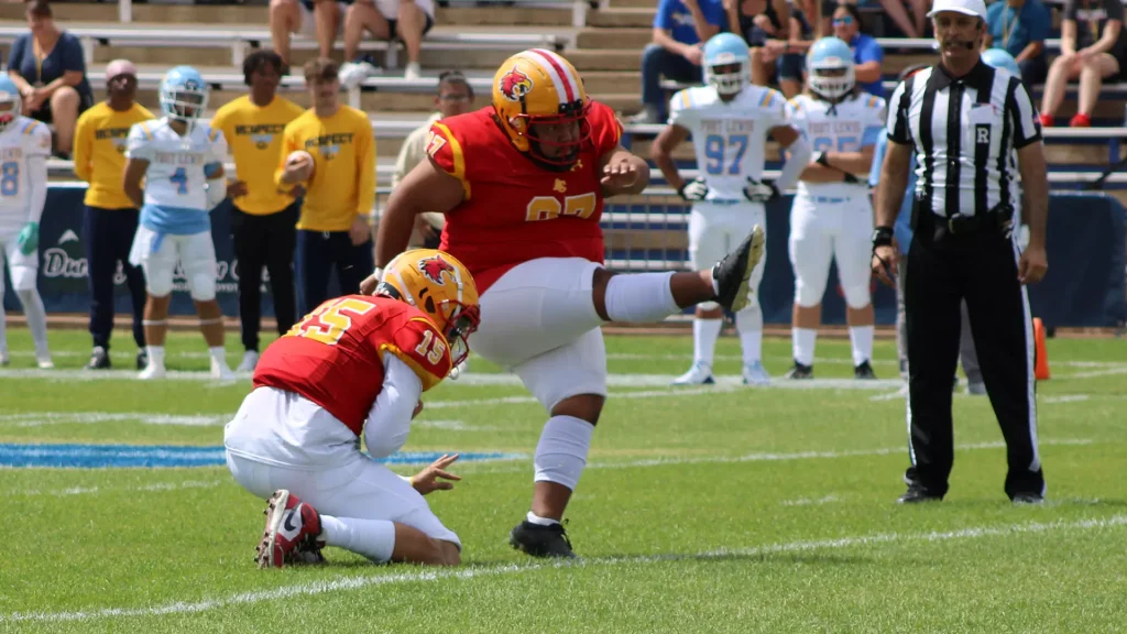 Football players in red and gold collide on a grassy field, with a referee nearby and teammates on the sideline in the background.