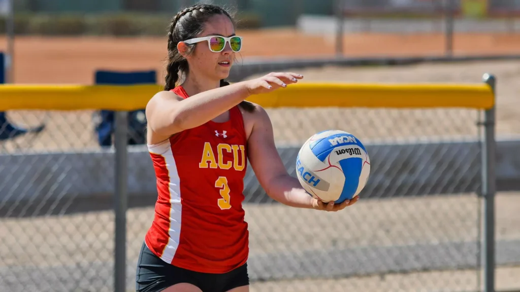 Female volleyball player in a red ACU jersey passes a ball on an outdoor court, wearing white sunglasses.