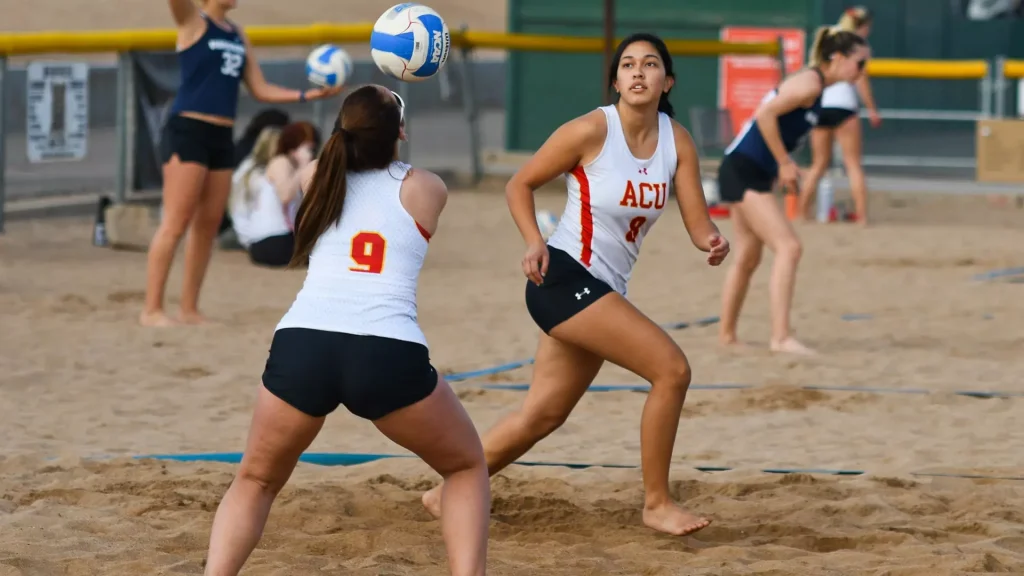 Two female beach volleyball players in white ACU jerseys chase a ball on a sandy court as it sails overhead.