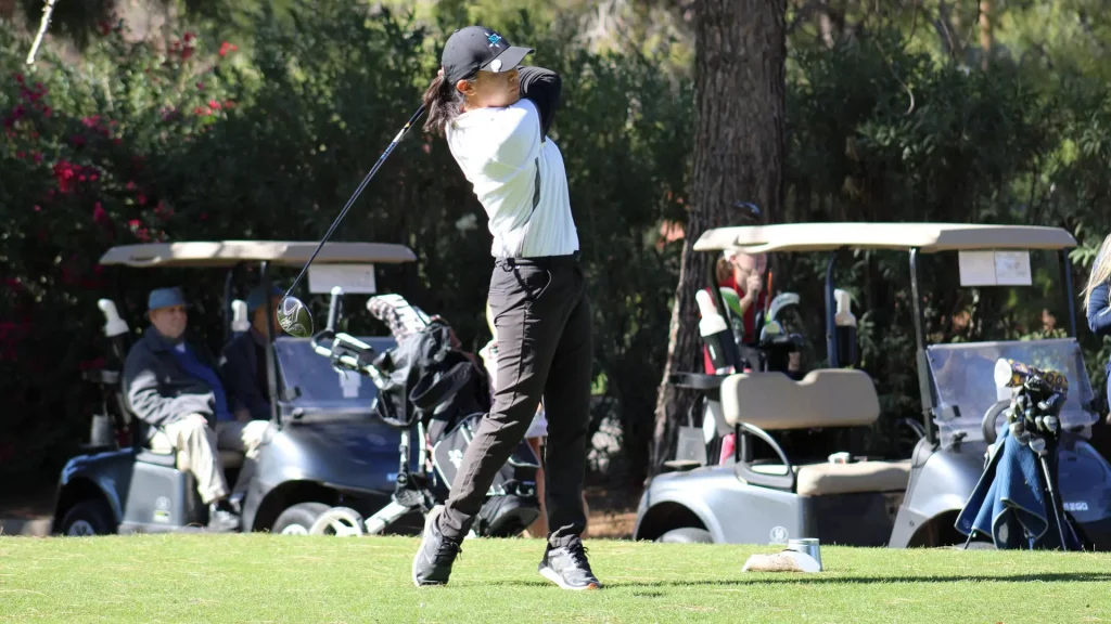 Woman golfer mid-swing on a sunny golf course, with golf carts and onlookers in the background.