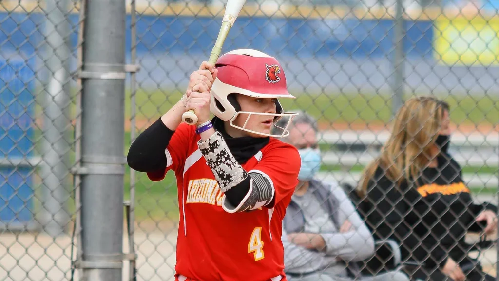 Youth baseball batter in a red jersey with number 4, gripping a bat and preparing to swing behind a chain-link fence