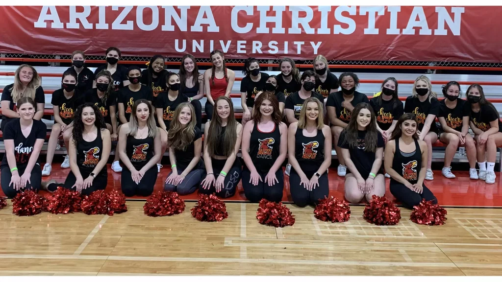 Large group of ACU cheerleaders in black uniforms posing on gym bleachers with a red 'Arizona Christian University' banner above them in the background, red pom-poms on the floor in front.