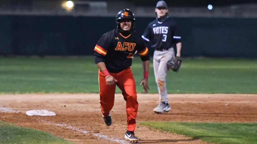 Baseball player in black and orange ACU uniform runs toward first base on a dusty infield, with a fielder in dark blue behind him.