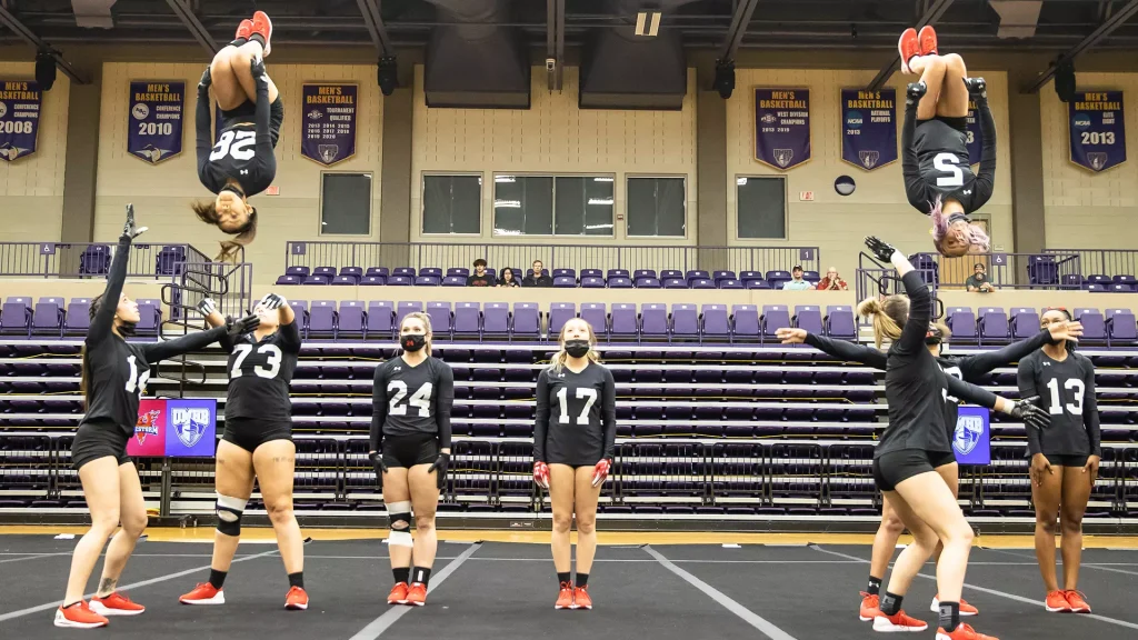 Girls in black uniforms with red shoes stand on a gym mat as teammates perform flips overhead, purple bleachers in the background.