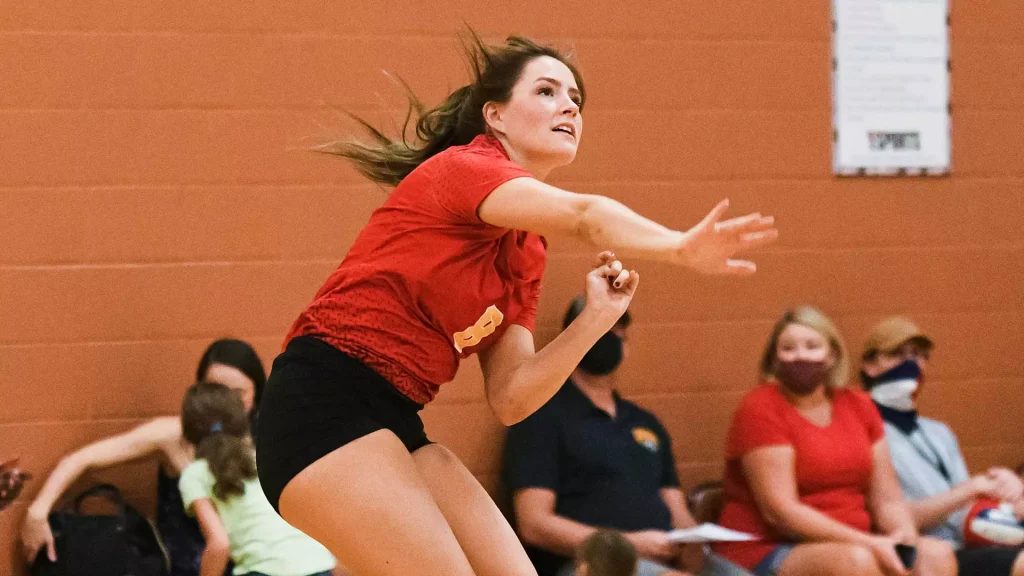 Female volleyball player in a red shirt and black shorts leaps forward with arms extended, mid-game in a gym as spectators watch from the benches against an orange wall.