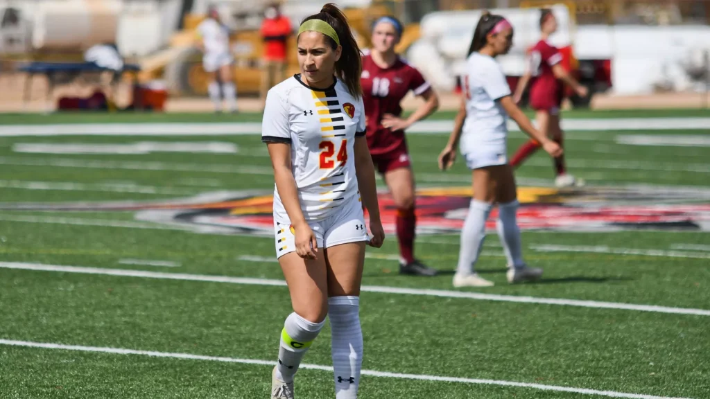 Female soccer player in white kit with number 24 walks on a green turf field during a daytime match, teammates in maroon and other players in the background.