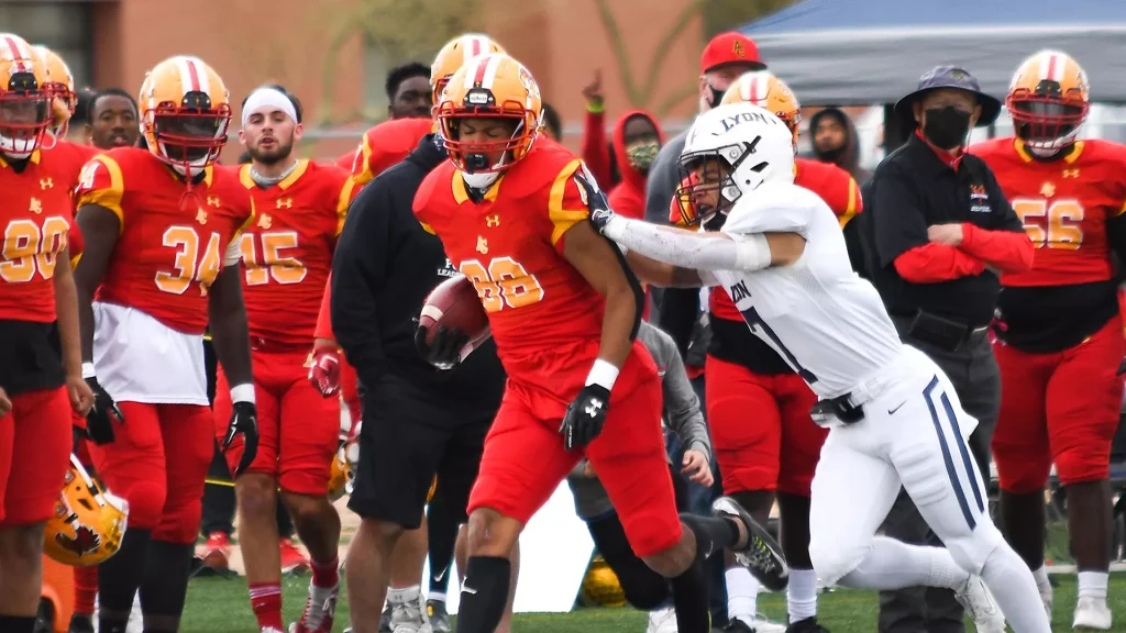 American football ball carrier in red jersey runs with the ball as a white-team defender reaches to tackle him.