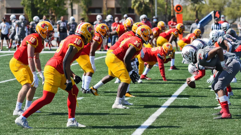 Football players in red and yellow uniforms line up at the line of scrimmage on a sunny field, ready for the snap.