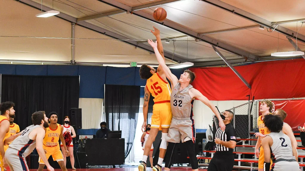 Two basketball players leap for the ball in a gym, one in orange (#45) and the other in gray, as teammates and spectators watch nearby.