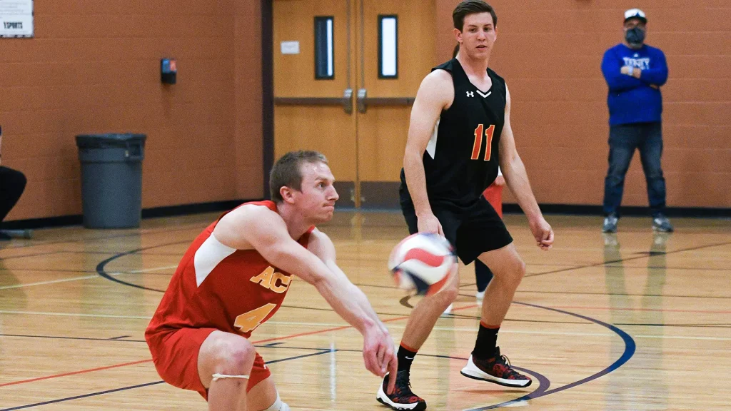 Two male volleyball players chase a ball on an indoor court; red jersey with #4 on the left, black jersey #11 on the right, spectator in blue in background.