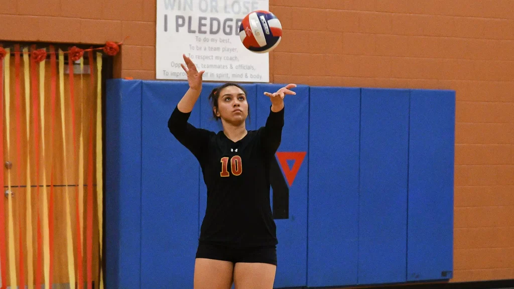 Female volleyball player in black uniform with number 10 serving a ball in a gym, with blue mat and orange wall behind.