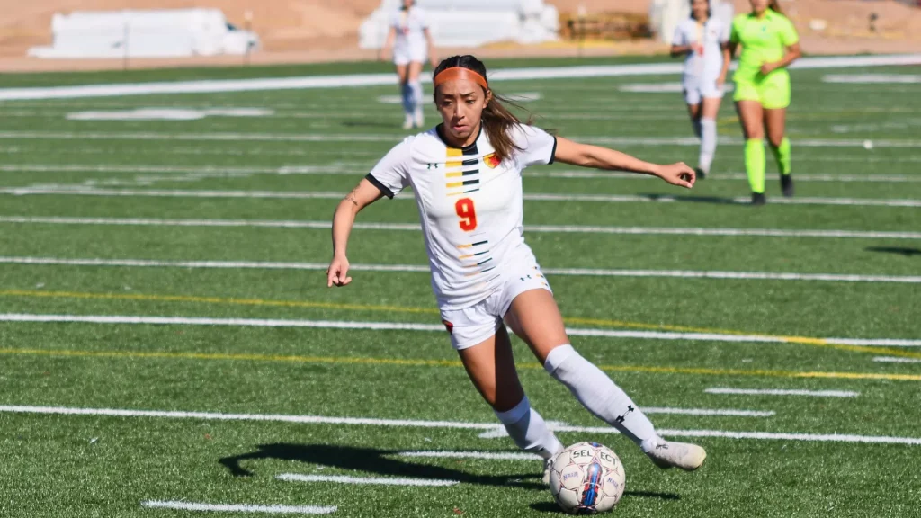 Female soccer player in white jersey with number 9 dribbles the ball on a turf field, orange headband in action.