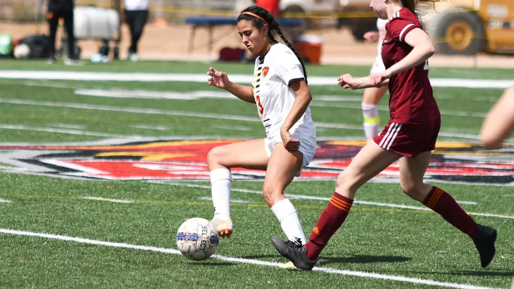 Two female soccer players compete for the ball on a green turf field; white uniform vs maroon uniforms in daytime action.