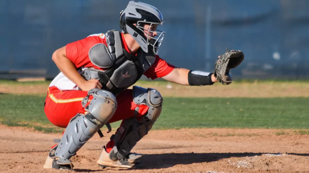 Baseball catcher in red uniform crouches behind home plate, glove extended for a play