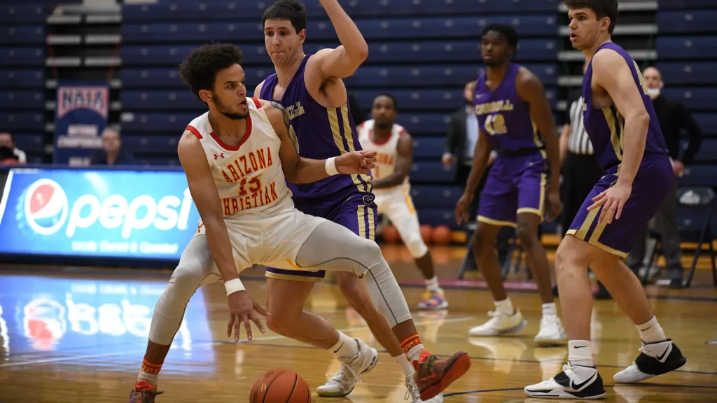 Arizona Christian player in white (#13) dribbles the ball as a purple-clad defender reaches in.