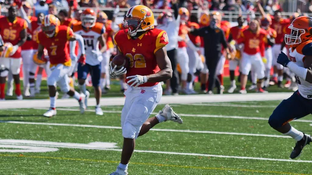 Running back in a red jersey with a yellow helmet carries the football while sprinting on a green field, crowd behind him.