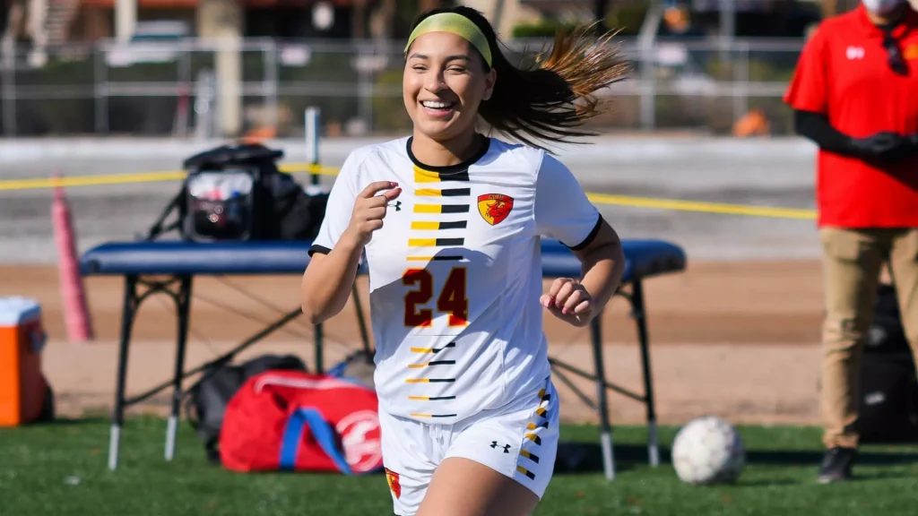 Female soccer player in a white kit with number 24 running on a sunny field, wearing a green headband.