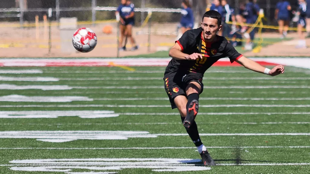 Soccer player in a black kit with red and yellow accents kicking a red-and-white ball on a green turf field, with spectators in the blurred background.
