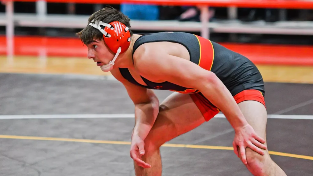 Wrestler in red and black singlet crouched on a wrestling mat, wearing red headgear and ready to start a match.
