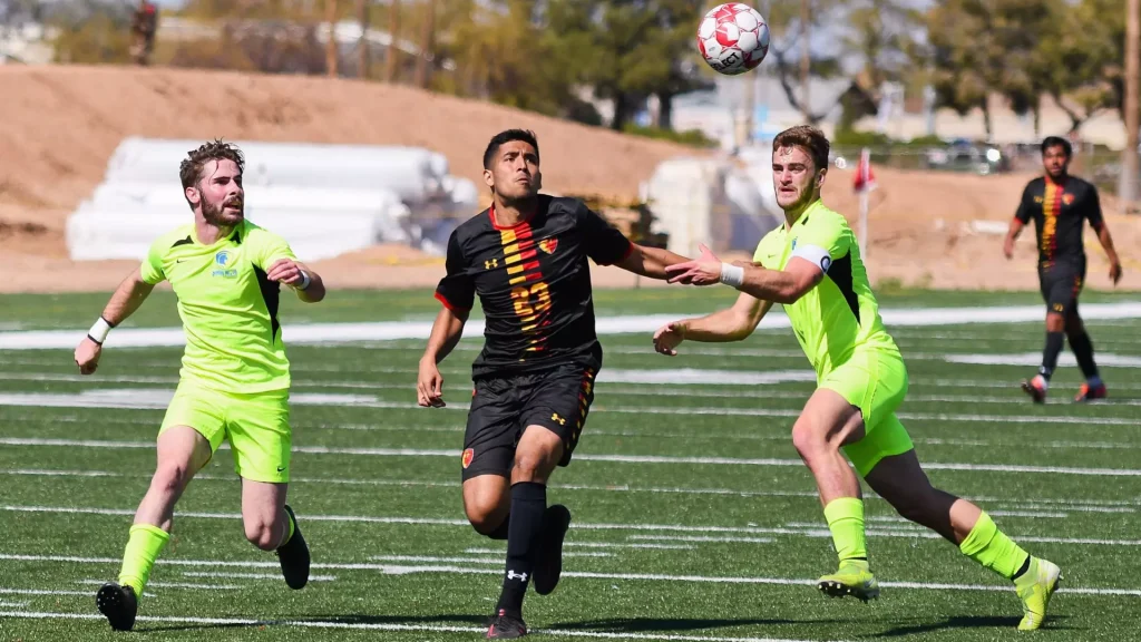 Soccer players chase a ball in midair on a green turf field, two wearing neon yellow uniforms and one in black with red accents.