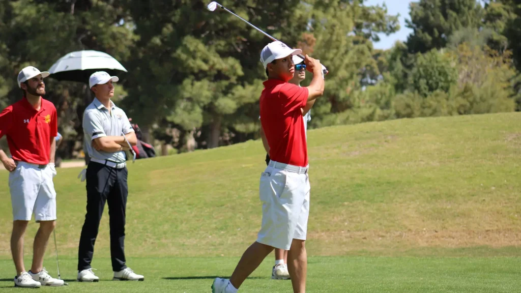 Golfer in a red shirt and white cap mid-swing on a sunny golf course, with teammates watching in the background.