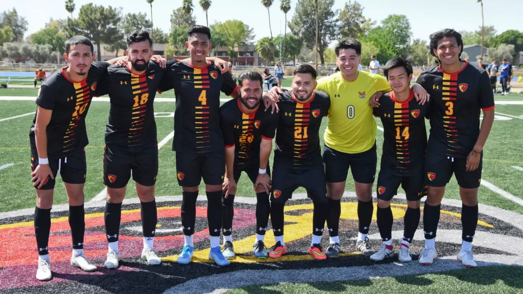 A group of eight soccer players in black uniforms with yellow-red stripes pose together on a field, arms around each other, smiling.