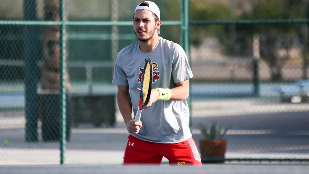 Male tennis player in a gray shirt and red shorts holding a racket on an outdoor court with a chain-link fence in the background.