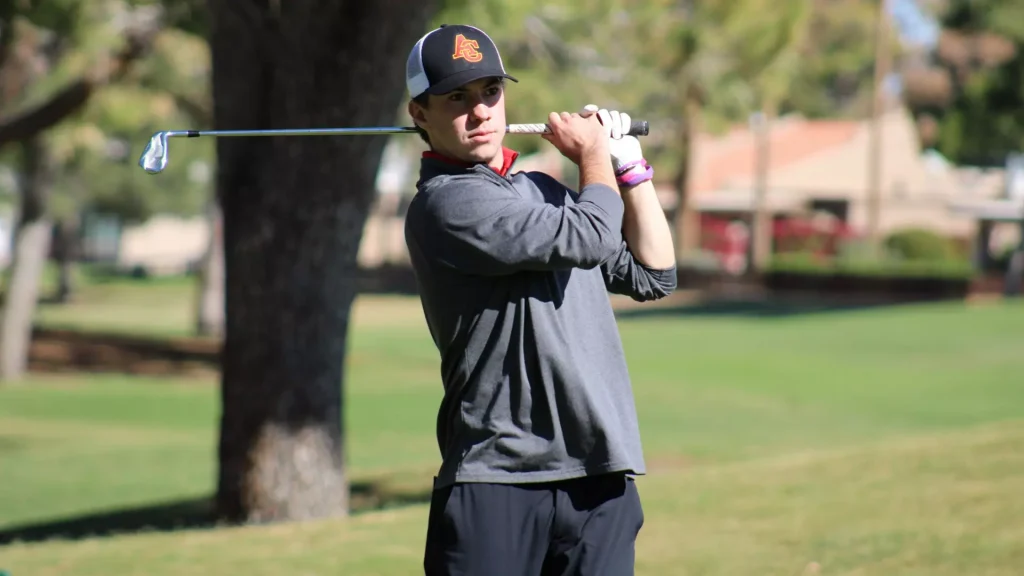 Man in a dark long-sleeve shirt and cap mid-swing with a golf club on a sunny golf course.